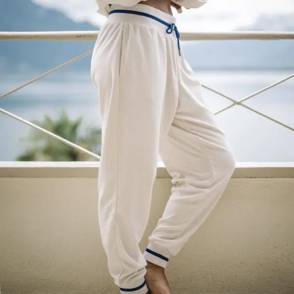 A person stands barefoot on a balcony in Vintage Trousers Terry Cloth with cream color and blue trim. The blurred background highlights water, mountains, and greenery.