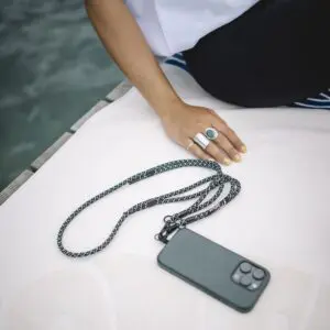 A person rests their hand on a white surface next to a smartphone attached to a Mobile Strap, Lakeside lanyard. They wear black pants, a white shirt, and silver rings featuring turquoise and green stone accents.