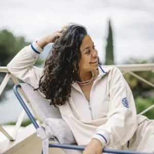 A woman with curly hair, wearing the unisex Terry Cloth zip-up trucker collar Vintage Sweat, sits outdoors on a chair, smiling and looking to the side with greenery and a cloudy sky in the background.