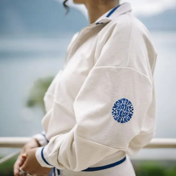 A person wears the unisex vintage Terry Cloth zip-up trucker collar Sweat in cream with blue trim and a circular blue NOT TRACK ATHLETIC logo on the sleeve, standing outdoors with a blurred lake and mountains behind.