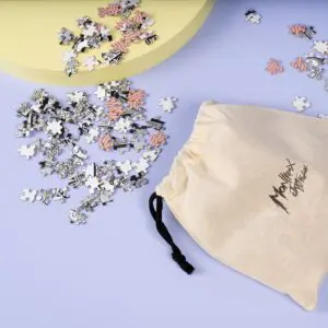 A small drawstring cloth bag lies on a light purple surface with scattered black, white, and pink 1000 Piece Rylsee Puzzle pieces nearby. A light yellow platform appears in the upper left corner.