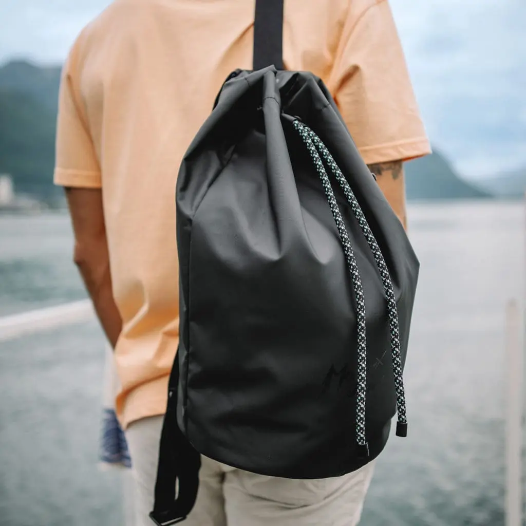 Vêtue d'une chemise orange clair et d'un pantalon beige, une personne se tient au bord de l'eau avec le sac Marin, Lakeside - un grand sac à cordon noir - en bandoulière. En arrière-plan, des montagnes et un ciel nuageux illustrent l'ambiance du festival de Montreux.