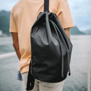 Wearing a peach t-shirt, a person carries the Lakeside Sailing Bag with a rope-style strap, standing outdoors near water and mountains, capturing the adventurous spirit of the Montreux Festival.