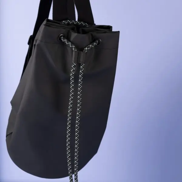 The Lakeside Sailing Bag, featuring black and white rope straps and a smooth matte finish, stands against a light purple background. Its minimalist design embodies the Montreux Festival’s spirit.