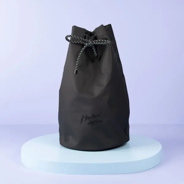 The Lakeside Sailing Bag in black, with a rope closure and subtle Montreux Festival logo, stands upright on a light blue circular platform set against a pale purple background—combining style and function.