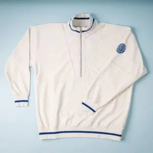 The Sweat unisex Terry Cloth zip-up trucker collar, Vintage, is a cream-colored pullover with a blue trucker collar, blue trim on the cuffs and hem, partial front zipper, and circular blue emblem on the left sleeve, shown flat on a light blue background.