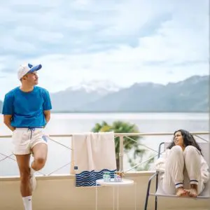 A man in the Vintage unisex Terry Cloth zip-up trucker collar sweatshirt and white shorts leans on a balcony, while a woman in light clothes sits nearby. A scenic lake and mountains stretch out under a cloudy sky behind them.