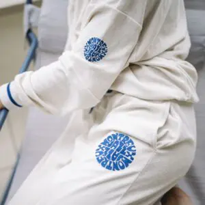A person sits on a cushioned chair wearing Vintage Trousers Terry Cloth and a matching cream tracksuit featuring blue circular "MONTEREY ALL DAY" text designs on the sleeve and pant leg.