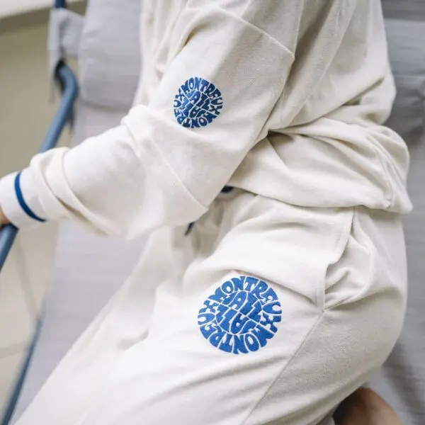 A person sits on a cushioned chair wearing Vintage Trousers Terry Cloth and a matching cream tracksuit featuring blue circular "MONTEREY ALL DAY" text designs on the sleeve and pant leg.