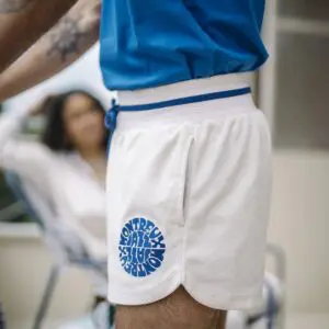 At an outdoor festival, a person wears the Short Terry Cloth, Vintage—white athletic shorts featuring a blue drawstring and a circular blue text graphic on the left leg. Another person is seen blurred in the background.