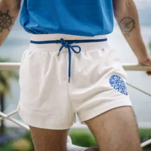 La personne porte une chemise bleue et un short en tissu éponge, Vintage, avec un cordon bleu et un motif circulaire bleu sur la jambe. Des tatouages nautiques ornent leurs bras, sur un fond festif de verdure et d'eau.