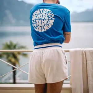 A person stands on a balcony with a lake and mountain view, wearing a blue T-shirt and the Short Terry Cloth, Vintage shorts with blue trim. A white towel hangs over the balcony railing.