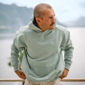 Un homme au crâne rasé et à la moustache porte un Sweatshirt brodé Montreux Jazz, Aloe, debout en plein air et regardant sur le côté, avec des montagnes et de l'eau floues créant un arrière-plan pittoresque et naturel.