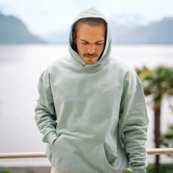 A man stands outdoors in an Aloe Sweatshirt Montreux Jazz embroidered, hood up and hands in pockets, gazing down. Behind him, water, mountains, and palm trees complete the serene scene.