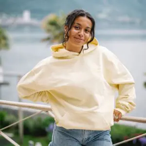 A woman stands outdoors by a railing, smiling with one hand on her hip, wearing blue jeans and a Sweatshirt Montreux Jazz embroidered, Butter. A lake and palm trees are visible in the blurred background.