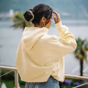 A woman with dark hair in a bun wears a Butter Sweatshirt Montreux Jazz embroidered and jeans, standing on a balcony overlooking water, palm trees, and distant mountains, turned away and shading her eyes with her hand.