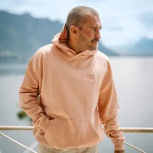 A man stands outdoors with his hands in the pockets of a Peach Sweatshirt Montreux Jazz embroidered, gazing to the side. A serene lake and mountains appear softly blurred in the background.