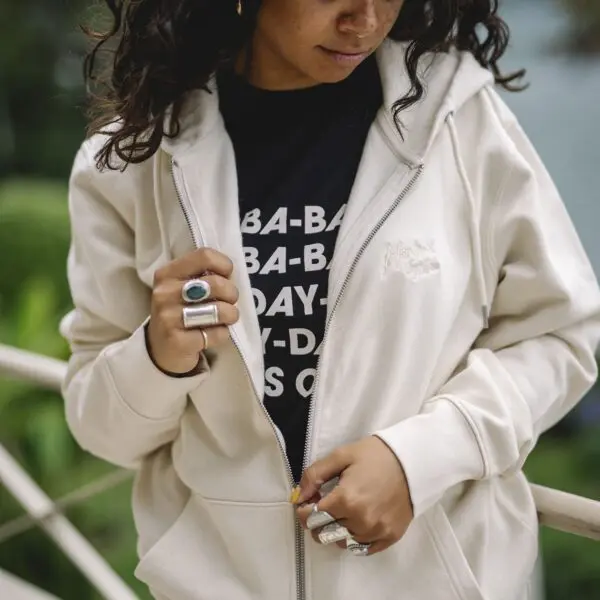 A person with curly hair stands outdoors near greenery, wearing the Embroidered zip-up hoody in Natural over a black graphic tee with white text, accessorized with large silver rings.