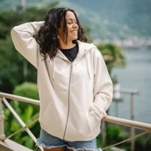 A smiling woman with curly hair poses outdoors in front of water and green hills, wearing the Natural embroidered zip-up hoody and denim shorts, with one hand behind her head.