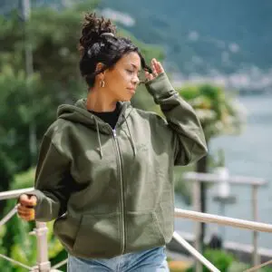 Une femme portant un Sweat à capuche zippé brodé, un Khaki et un jean se tient en plein air au bord d'un lac, se protégeant du soleil, avec de la verdure et des montagnes en arrière-plan.