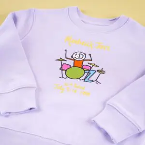 Un sweat-shirt lavande présente un dessin coloré et enfantin d'un batteur souriant à une batterie, avec les textes "Montreux Jazz" et "32nd Festival July 3-18 1998" écrits au-dessus et au-dessous de l'illustration.