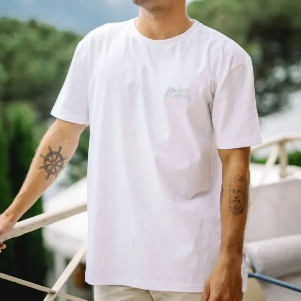 Portant le T-Shirt Montreux Jazz, Aloe, une personne se tient à l'extérieur, la tête partiellement hors du cadre. Des tatouages couvrent les deux avant-bras, tandis qu'une verdure douce et floue et des touches d'aloès créent une ambiance de fond.