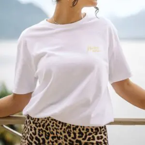 A person wears a white T-Shirt Montreux Jazz, Butter and a leopard print skirt while standing outdoors, leaning on a railing with a blurred scenic background of mountains and water.