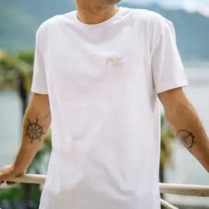 A person wearing a T-Shirt Montreux Jazz, Peach with a small peach logo on the chest stands outdoors by a railing. Palm trees and water are blurred in the background, and tattoos are visible on both forearms.