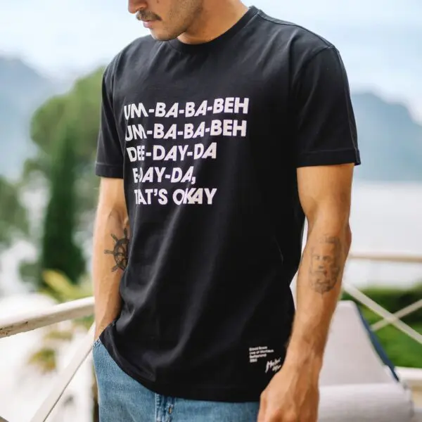 A man stands outdoors in a black T-Shirt Under Pressure featuring bold white text, reflecting the Montreux Jazz spirit. Blurred greenery and a railing are visible in the background.