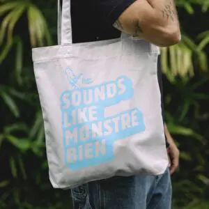 A person holds the Tote Bag Rylsee, Blue, featuring light blue "SOUNDS LIKE MONSTRE BIEN" text and a guitar-playing monster. Lush green plants are blurred in the background, creating a fresh Montreux Blue ambiance.