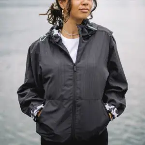 A woman stands by a lake, hands in her pockets, wearing the Lakeside Water-Repellent Hooded Jacket with camo-print inner cuffs—perfect for festivals—her loose waves and hoop earrings completing the look.