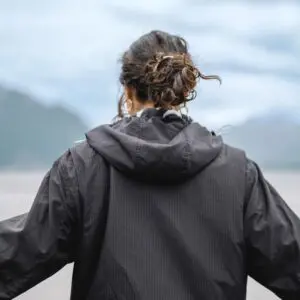 A person with curly brown hair in a loose bun wears the Water-Repellent Hooded Jacket, Lakeside, standing outdoors against a blurred natural landscape and cloudy sky—ready for any festival weather.