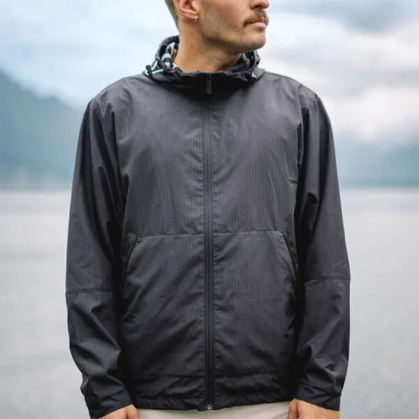 A man in the Water-Repellent Hooded Jacket, Lakeside, stands by the water with mountains behind him—ideal festival outerwear. His head is turned slightly to the side.