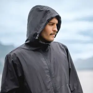 A man in a Lakeside Water-Repellent Hooded Jacket stands outdoors at a festival, gazing to the side. The blurred background reveals a cloudy sky and faint mountain outlines.