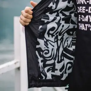 A person holds open a Water-Repellent Hooded Jacket, Lakeside, revealing its bold black and white abstract patterned lining. They wear a black shirt with white text, ideal for festival style against a blurred outdoor background.