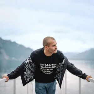 A man with a shaved head and mustache leans on a white railing by a lake and mountains under a cloudy sky, wearing the Lakeside Water-Repellent Hooded Jacket—a perfect choice for a festival outing.