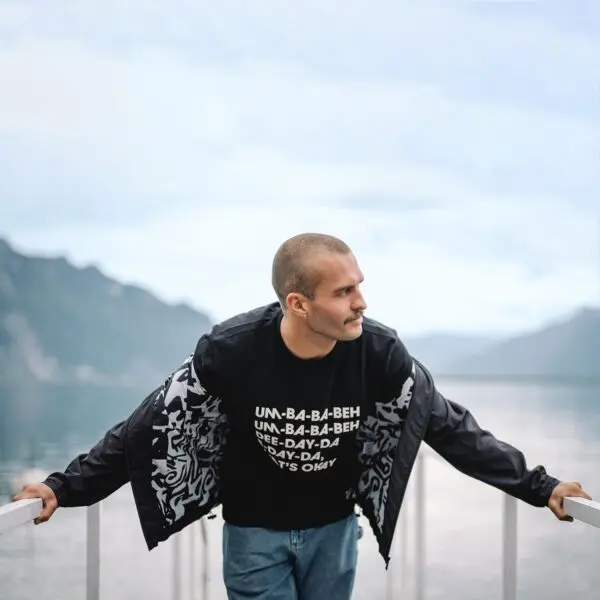 A man with a shaved head and mustache leans on a white railing by a lake and mountains under a cloudy sky, wearing the Lakeside Water-Repellent Hooded Jacket—a perfect choice for a festival outing.