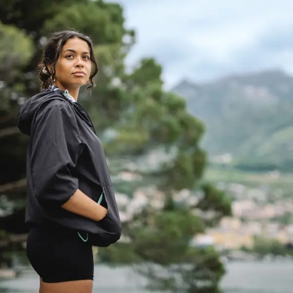 Wearing the Lakeside Water-Repellent Hooded Jacket, a woman stands outdoors among trees and mountains, gazing thoughtfully to the side on a cloudy day, ready for any festival adventure.