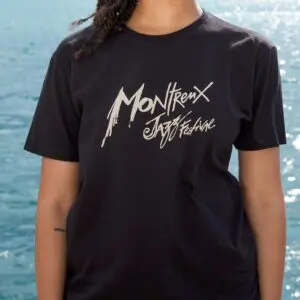 A person in a T-Shirt Montreux Jazz Festival, Black, stands before blue water. Only their upper torso and part of an arm with a small tattoo are visible.