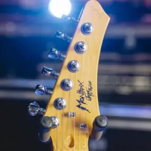 Close-up of a São Paulo, Guitar Collection, Limited Series headstock with tuning pegs, strings, and signature, set against a blurred background with bright stage lights.