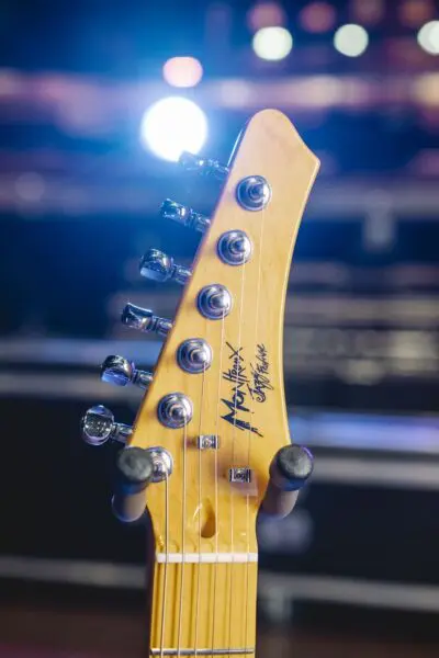 Close-up of a São Paulo, Guitar Collection, Limited Series headstock with tuning pegs, strings, and signature, set against a blurred background with bright stage lights.