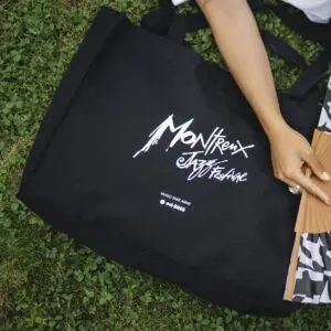 A Shopping Bag, Montreux Jazz, with white Montreux Jazz Festival text, sits on green grass next to an arm holding a paper fan.