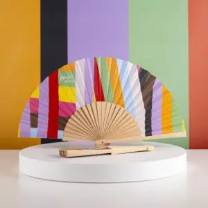L'Éventail Lakwena, un éventail à main avec des rayures multicolores et un manche en bois, est présenté ouvert sur un support blanc à côté de l'éventail fermé assorti, sur fond de rayures orange, noires, vertes et pêche.
