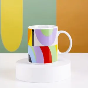 The Mug Lakwena is a white ceramic mug with a vibrant abstract pattern, displayed on a white pedestal with a multicolored geometric background inspired by Montreux Jazz festival vibes.