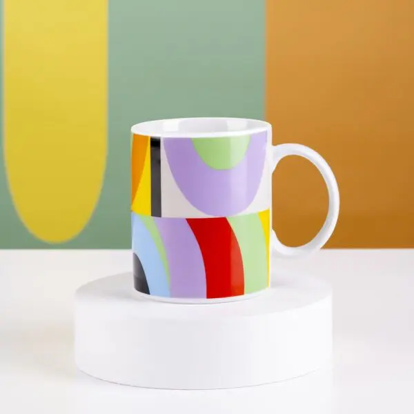 The Mug Lakwena is a white ceramic mug with a vibrant abstract pattern, displayed on a white pedestal with a multicolored geometric background inspired by Montreux Jazz festival vibes.