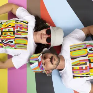 Deux personnes allongées sur une surface rayée colorée, portant toutes deux des t-shirts blancs avec des textes multicolores vibrants et des chapeaux assortis ; l'une a des lunettes de soleil rondes et des cheveux longs, l'autre une moustache et une courte barbe.