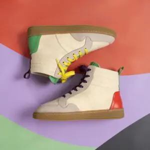 Two Lakeside Wooler Sneakers by Baabuk, Limited Edition, with beige suede uppers, green and red heel details, brown soles, and mismatched laces are showcased on an abstract Montreux Festival-inspired background.