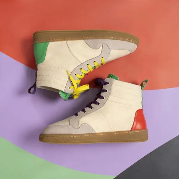 Two Lakeside Wooler Sneakers by Baabuk, Limited Edition, with beige suede uppers, green and red heel details, brown soles, and mismatched laces are showcased on an abstract Montreux Festival-inspired background.