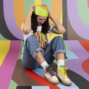 A person with long curly hair in a rainbow headscarf, rolled-up jeans, and Limited Edition Lakeside Wooler Sneakers by Baabuk sits cross-legged on a colorful striped backdrop, smiling and soaking in the Montreux Festival vibe.