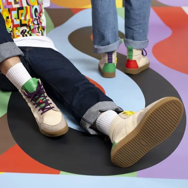 At the Montreux Festival, two people in rolled-up jeans show off their Lakeside Wooler Sneakers by Baabuk, Limited Edition. Only their lower legs and feet are visible on a bold, abstract-patterned floor—the sneakers’ bright accents stand out.